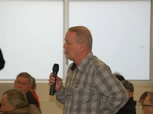 In this pictures so many people are sitting on a chair and one person is standing and he is holding...