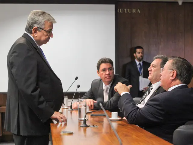 This picture shows few people seated on chairs and couple of them standing and we see a man holding...